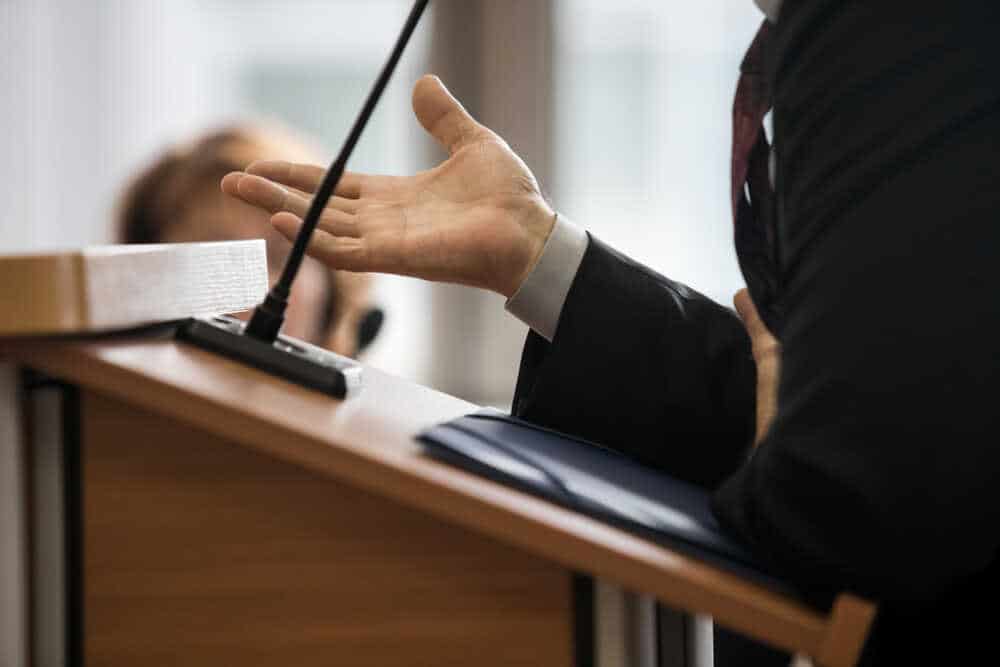 A person in a suit is speaking at a podium with one hand raised, gesturing mid-speech. A microphone is mounted on the podium, likely addressing issues relevant to their role as a Criminal Defense Attorney in Harford County. Another attendee is slightly visible in the background, out of focus.