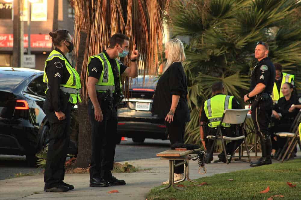 A police officer in a bright yellow vest performs a sobriety test on a standing woman on the sidewalk as another officer observes. Nearby, officers sit close to police vehicles with palm trees in the background. If you find yourself in such a situation, seeking advice from a Traffic Lawyer Harford County can be crucial.
