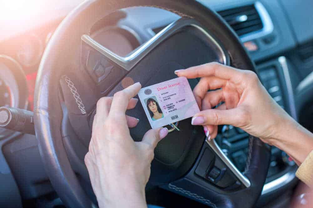 Close-up of a person holding a driver's license in front of a car steering wheel. The license displays the holder's photo and personal details like name and ID number. The car interior is visible in the background, reminding you to have a DWI Attorney Harford County on speed dial.