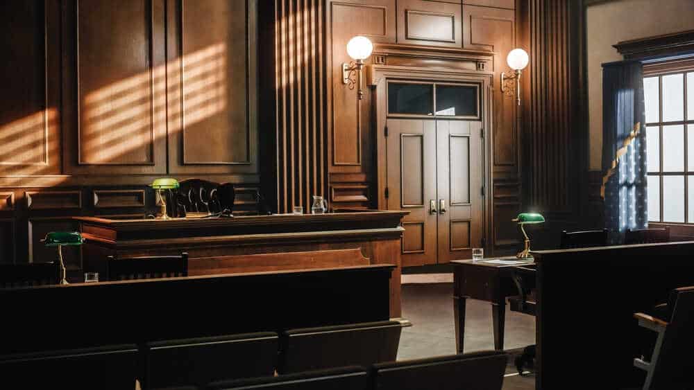 An empty courtroom with wooden furnishings, including a judge's bench, witness stand, and several rows of benches. Two green desk lamps are visible, with sunlight streaming through partially closed blinds, casting shadows on the walls and floor—the very setting where a Criminal Defense Attorney Cecil County might fiercely advocate for justice.