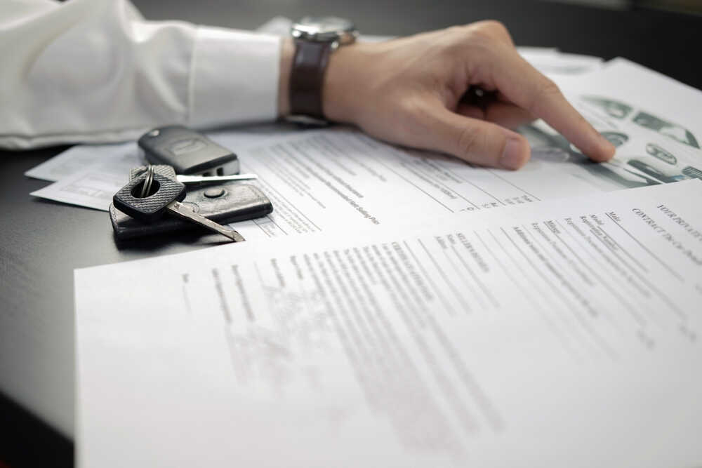 A person's hand is pointing at a car sales contract on a black table. Car keys are placed beside the contract, along with other documents in the background. The person, potentially consulting a DWI Attorney Cecil County for legal advice, is wearing a white shirt and a watch.