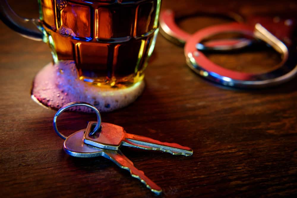 A close-up shot shows a beer mug with foam spilling onto a wooden table, alongside car keys. In the background, out-of-focus handcuffs are visible, suggesting a theme related to drinking and driving. The scene is dimly lit with a warm, amber glow—a stark reminder to seek guidance from a Traffic Lawyer in Cecil County.