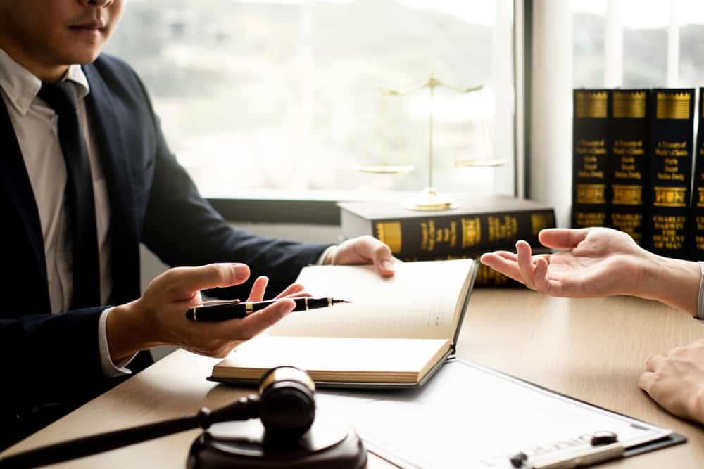 Two people discuss legal matters in an office setting. One person in a suit, possibly a Criminal Defense Attorney from Harford County, points to a book while holding a pen, and the other person's hand gestures. A gavel, legal books, and scales of justice adorn the desk.